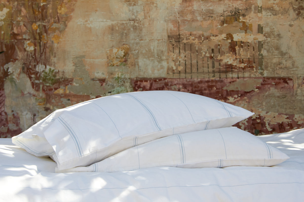 Two white pillows with blue stripes on a bed, rustic wall behind.